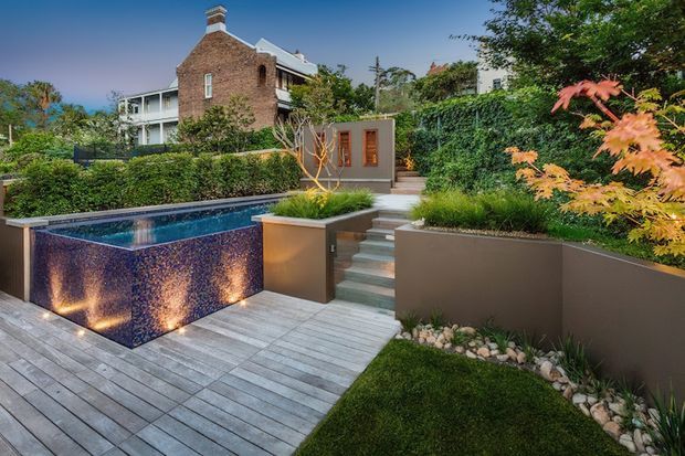 Landscaped residential backyard with a raised swimming pool clad in a dark blue-purple textured finish, timber decking, stepped garden terraces, hedges, ornamental planting, and a small timber door or gate. A multi-storey brick-and-white house is visible beyond the garden.
