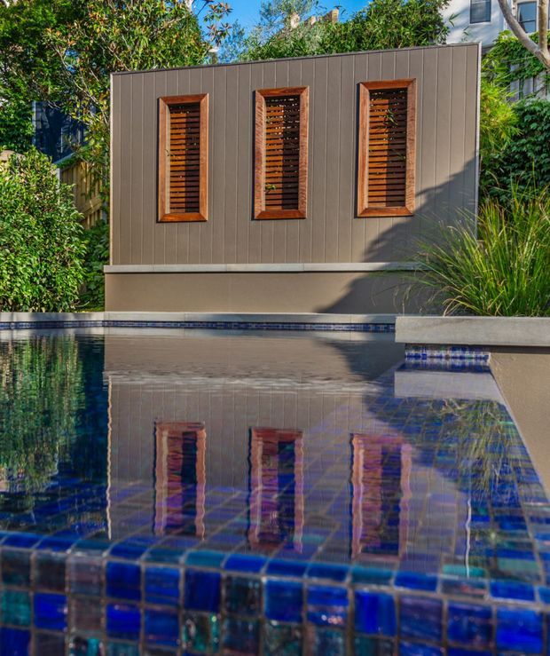 A close-up exterior view of a modern landscaped pool courtyard. The rectangular swimming pool has deep blue mosaic-tiled edging and reflects a dark vertical-panel wall with three timber-framed louvred openings. Lush planting surrounds the pool, with a raised concrete planter at right and a contemporary building partially visible behind the greenery.