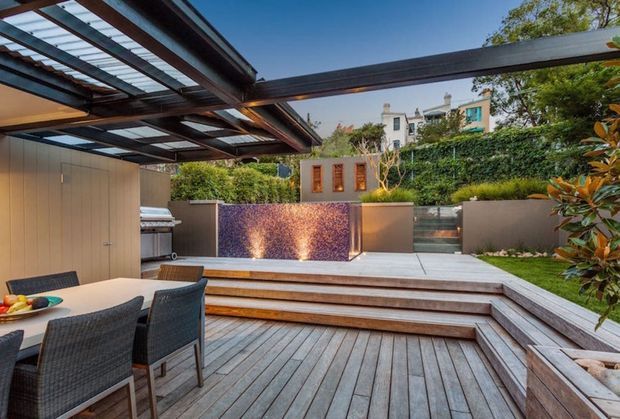 A landscaped outdoor entertaining area viewed from a covered dining space, featuring a timber deck with multiple steps, an outdoor dining table, a built-in barbecue, a tiled water feature or pool wall, raised garden beds, hedging, and a steel-framed translucent roof structure. The setting is contemporary and warmly lit with integrated feature lighting.