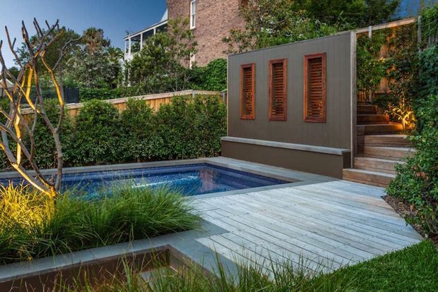 A landscaped residential courtyard with a compact rectangular swimming pool, pale timber decking, planted garden beds, and a dark rendered feature wall with three timber louvred panels. Steps lead upward through dense greenery toward a brick building, creating a private and layered outdoor setting.
