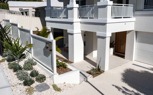 Bright exterior view of a contemporary two-storey residence featuring a white rendered facade, covered entry porch supported by square columns, upper-level balcony with white vertical balustrades, timber front door, attached garage, and a low-maintenance landscaped frontage with succulents and gravel.