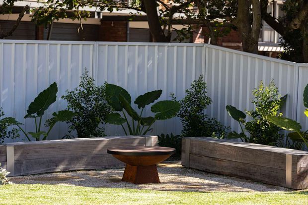 A landscaped outdoor courtyard or garden seating area with a low timber bench, a sculptural wooden pedestal table, raised timber garden beds, tropical foliage, gravel, lawn, and a white corrugated metal boundary fence. Brick and timber residential elements are partially visible beyond the fence.