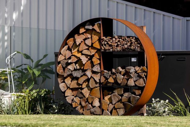 Outdoor garden area featuring a sculptural circular firewood storage rack filled with split logs, positioned against a corrugated metal-clad wall and dark cabinetry or screening. Low planting borders and lawn surround the feature, with strong sunlight creating a crisp, contemporary outdoor setting.