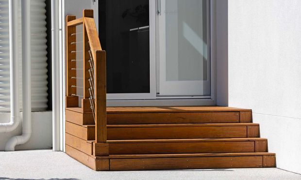 Exterior view of a compact set of warm timber steps leading to a raised entrance with a white-framed glazed door or window. The steps include solid timber risers and integrated side railings with thin metal cables. White rendered walls, visible external plumbing, and strong sunlight create a clean, functional facade detail.