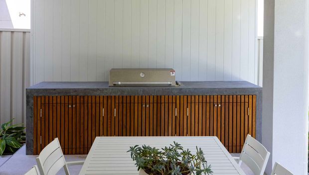 A covered outdoor entertaining area with a long built-in timber-slatted cabinet and integrated barbecue against a white vertical-panel wall. A white outdoor dining table with potted greenery and matching chairs occupies the foreground, creating a clean contemporary alfresco setting.