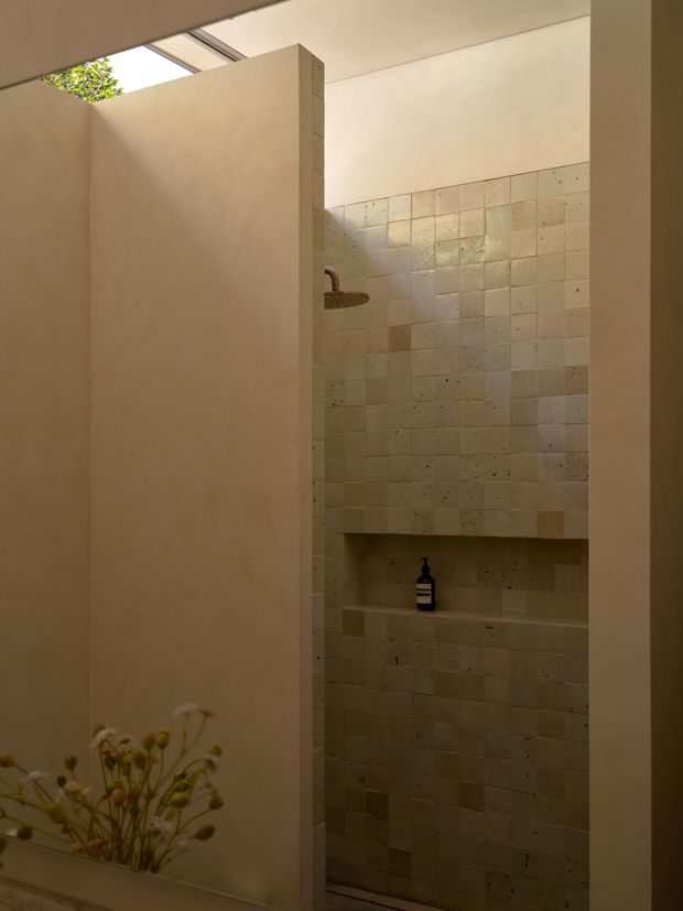 A warm, minimalist walk-in shower area viewed through a partial-height partition. The shower has a large round metal showerhead, a recessed tiled niche, and full-height small-format beige stone or ceramic tiles with subtle tonal variation. Smooth pale plaster walls and a skylight bring in soft natural light.
