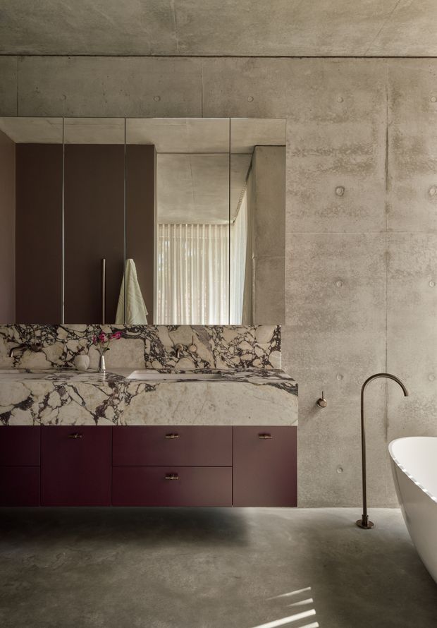 A sophisticated modern bathroom featuring a floating burgundy vanity with brass hardware, a dramatic veined marble benchtop and backsplash, a large wall-to-wall mirror, exposed concrete walls and floor, and a freestanding white bathtub with a dark arched floor-mounted tap. Natural light filters through vertical blinds, creating a refined, moody atmosphere.