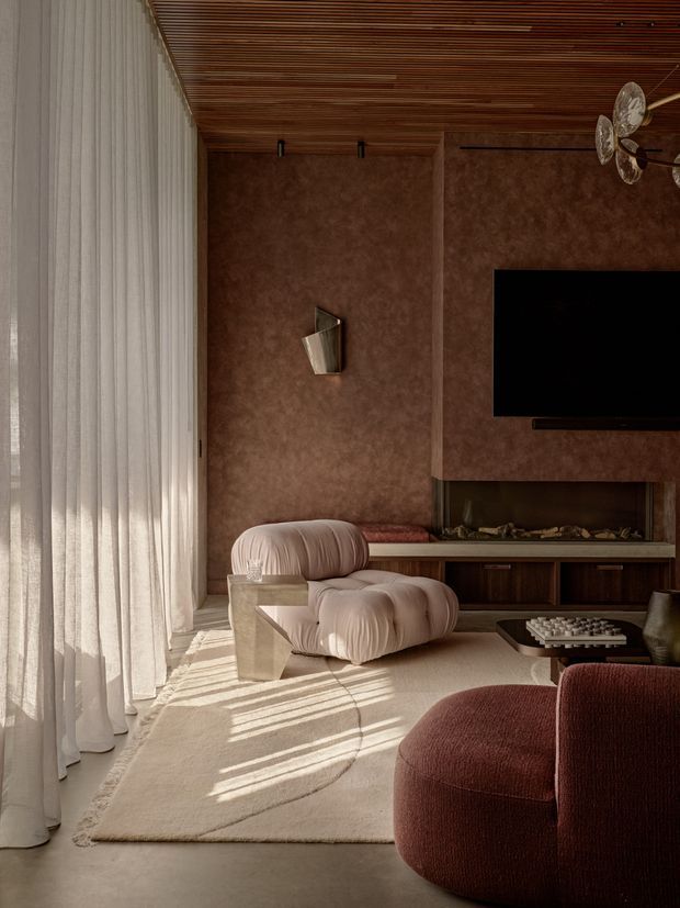 A warm, moody living room with floor-to-ceiling sheer curtains, a richly textured brown feature wall, and a slatted timber ceiling. The room includes a wall-mounted television with low dark timber joinery, a sculptural cream lounge chair, a deep burgundy upholstered sofa in the foreground, a pale textured rug, and a small dark coffee table. Soft sunlight creates strong linear shadows across the rug and floor, while a decorative wall sconce and pendant light add refined detail.