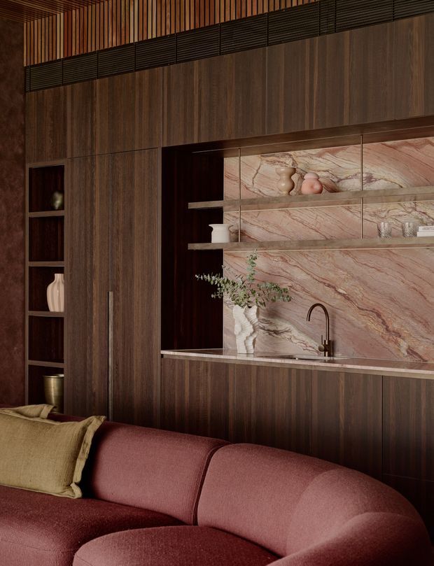 A richly detailed open-plan living area with a compact integrated kitchenette or bar behind a curved burgundy sofa. The room features dark vertical timber cabinetry and wall panelling, a dramatic veined pink-and-cream stone splashback and benchtop, open display shelving, a dark curved tap, and warm timber slatted ceiling detailing. The overall composition is warm, moody, and sophisticated.
