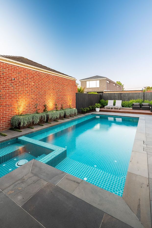 A bright residential backyard pool area with a large turquoise tiled swimming pool, integrated square spa, and broad grey stone paving. The pool is enclosed by a red brick wall and dark vertical timber fencing, with linear planting beds, outdoor lounge chairs, and a compact seating area along the far edge. Suburban homes and pitched roofs are visible beyond the fence under a clear sky.