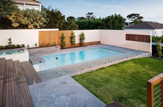 A landscaped residential backyard with a large rectangular swimming pool enclosed by frameless glass fencing. The pool is surrounded by dark timber decking, irregular grey stone paving, lawn, rendered boundary walls, and timber screening panels. A raised spa or shallow water feature sits at the left, with established planting around the perimeter.