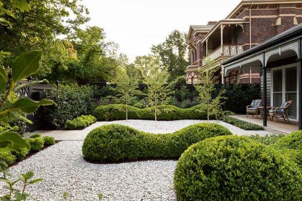 A formal landscaped courtyard garden beside a substantial brick residence. Rounded clipped hedges and small ornamental trees are arranged in geometric beds over a pale gravel ground plane, with a covered verandah and decorative timber detailing along the house.