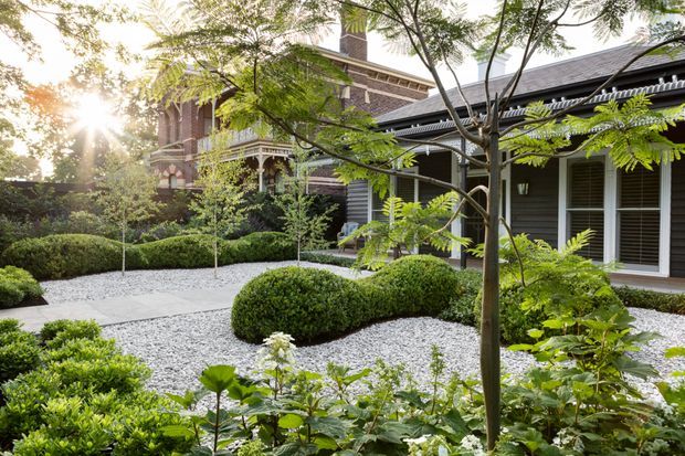 A landscaped courtyard garden beside a heritage-style brick building, featuring a white gravel ground plane, clipped rounded hedges, layered planting, slender trees, and a covered verandah with timber posts and detailed traditional trim. The composition combines a richly ornamented brick facade with a darker weatherboard-like wing and large glazed doors.