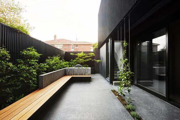 A contemporary narrow courtyard beside a dark, vertically screened building. The space features black metal cladding and glazing, a long built-in timber bench, concrete paving, raised planting beds, lush greenery, and a high privacy fence. A neighbouring brick roofline is visible beyond the courtyard.