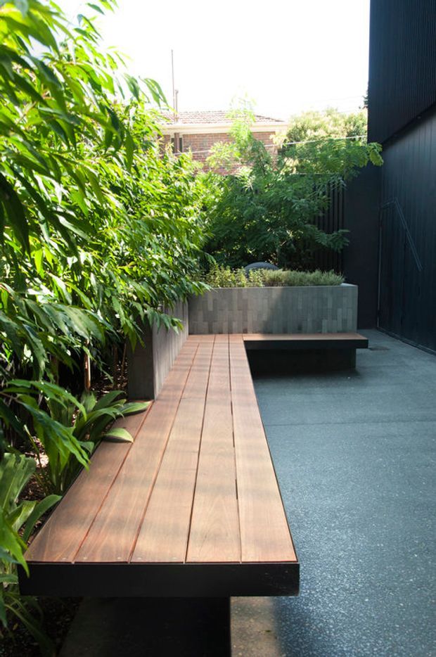 A narrow landscaped courtyard or side garden with a long raised timber bench/deck running along the planting bed. Dense green foliage borders the space, while a dark vertically clad wall and smooth grey paving create a contemporary outdoor retreat. A rendered masonry planter with integrated seating is visible at the far end, with a neighbouring brick building beyond.