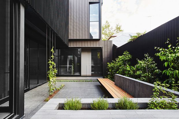 A contemporary dark-clad residential courtyard viewed from outdoors, featuring charcoal vertical wall cladding, extensive black-framed glazing, a paved courtyard, raised planted beds, lush greenery, and a built-in timber bench along a low masonry wall. The composition highlights a private, landscaped outdoor space enclosed by the surrounding building forms.