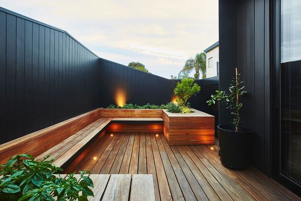 A compact, enclosed urban courtyard deck with warm timber decking and built-in timber bench seating arranged around a recessed, illuminated feature area. Tall dark vertical-panel fencing and matching dark cladding create privacy, while potted and planted greenery soften the space. Large dark-framed glazing is visible along the right side, and integrated low-level lighting adds a warm evening ambience.