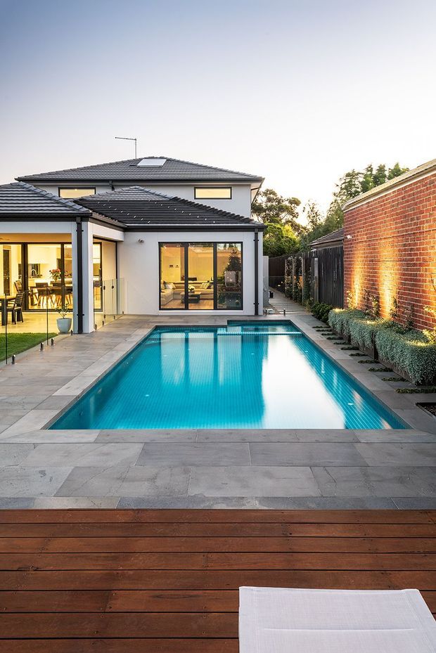 A contemporary suburban backyard viewed from a timber deck, featuring a rectangular turquoise swimming pool surrounded by large grey paving slabs. The two-storey white-rendered house has dark tiled hipped roofs, extensive black-framed glazing, and a small skylight. A tall red-brick boundary wall and landscaped screening line the right side of the pool.