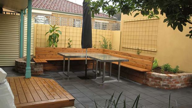 A compact enclosed courtyard or patio with built-in timber bench seating arranged around a long outdoor dining table and closed umbrella. The space has dark grey tiled paving, timber decking, brick-edged raised planters, cream rendered walls with grid detailing, and a tiled roof visible beyond the boundary wall. Greenery and a corrugated roller door are present at the edges.