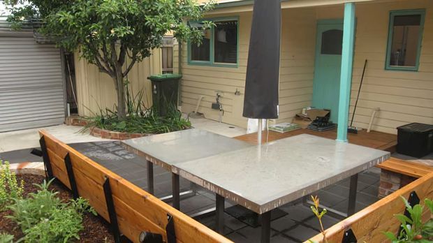A compact outdoor courtyard dining and entertaining area beside a single-storey weatherboard building. The space features a large built-in outdoor table with bench seating, a central dark suspended or hooded feature, paved flooring, a turquoise-painted entry door and posts, timber wall cladding, and surrounding garden planting.