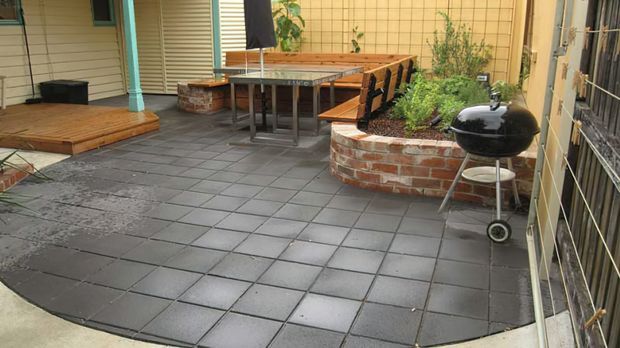 A paved courtyard-style outdoor entertaining area with large dark square pavers, timber decking at the edges, a built-in curved brick planter, outdoor dining furniture, and a barbecue. The space is enclosed by weatherboard walls and fencing, with greenery in raised planters.