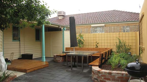 A compact enclosed courtyard entertaining area with a timber deck, outdoor dining table and bench seating, raised brick garden beds, potted plants, and a covered patio structure. The surrounding single-storey building has pale weatherboard walls, turquoise-painted posts, and a tiled pitched roof. A barbecue and closed outdoor umbrella complete the functional outdoor setting.