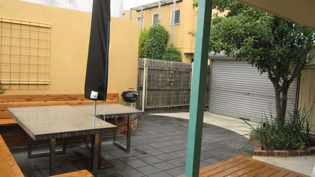 A compact paved courtyard entertaining area viewed from under a covered structure, with timber bench seating, a tiled outdoor dining table, a closed barbecue, raised garden beds and mature planting. A roller garage door and surrounding rendered walls frame the urban courtyard.
