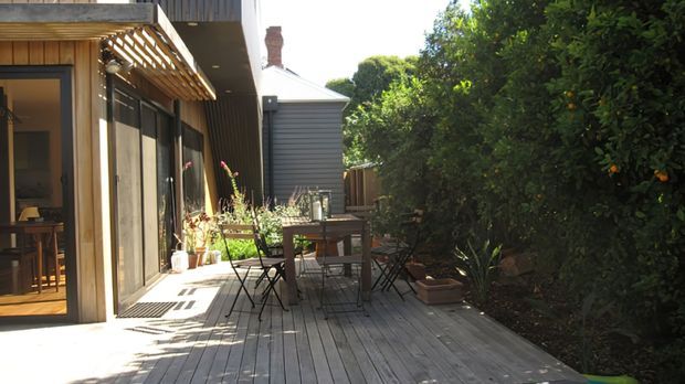 Sunlit timber deck beside a contemporary residential extension, with large glazed doors, a pergola-like timber screen, outdoor dining furniture, and dense garden planting along the boundary. A neighbouring weatherboard structure and roofline are visible beyond the deck.
