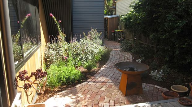 A narrow landscaped side courtyard beside a modern home, featuring a curved brick-paved walkway, planted garden beds, timber and dark horizontal wall cladding, and a small sculptural outdoor fire bowl or feature table. Dense greenery screens the boundary, creating a private garden passage.