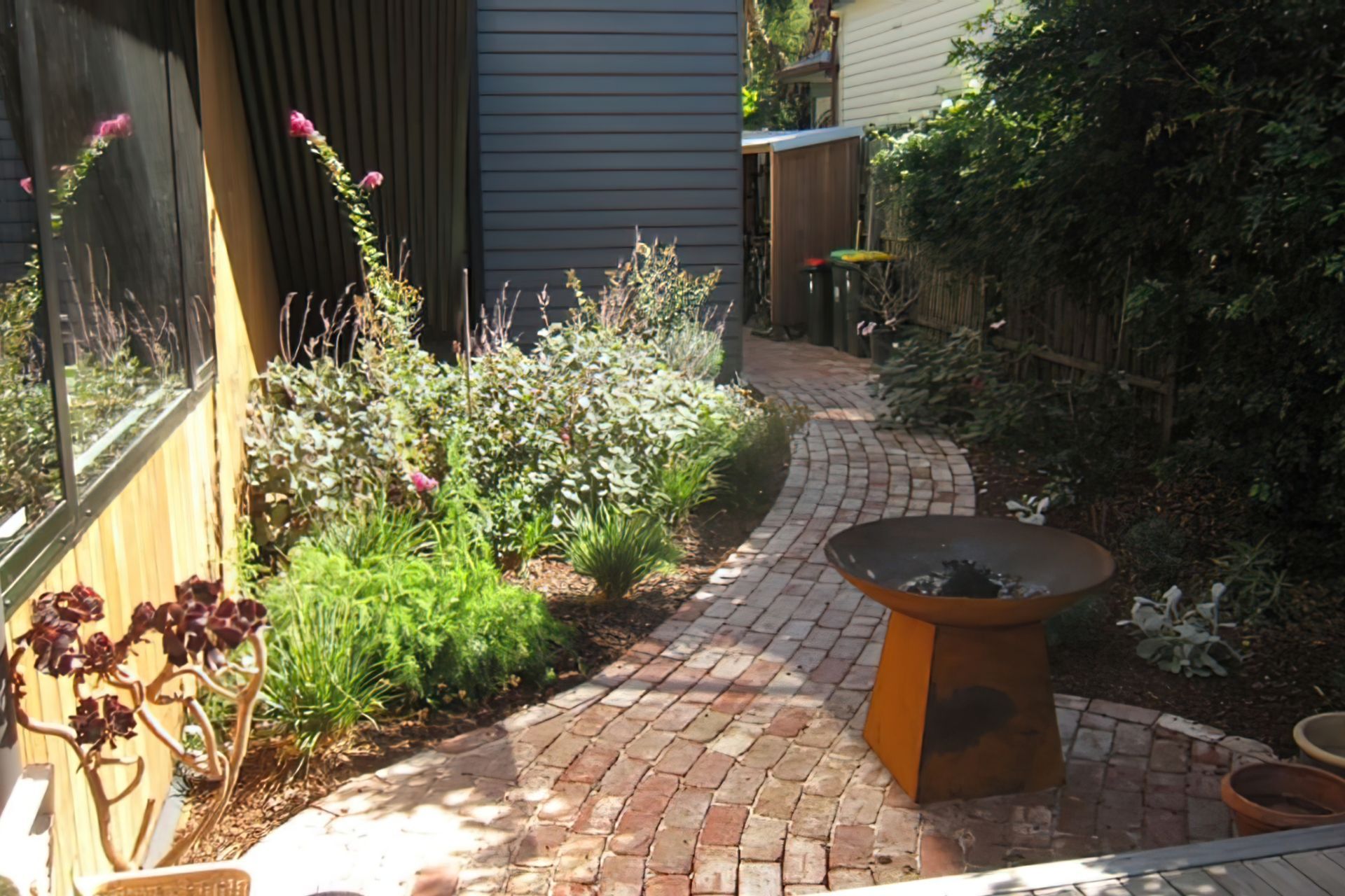 AFTER the garden design. A sinuous red brick path enters the deck at a small gathering node where a firepit is placed