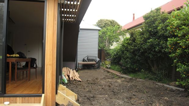 A compact residential backyard viewed from a timber-clad extension or covered outdoor area. The foreground is a freshly excavated or prepared soil area bordered by dense hedging, with a narrow timber path leading toward a small corrugated-metal shed or outbuilding. An open glazed doorway reveals a simple timber-floored interior and dining furniture. The image shows a modest suburban site with mixed timber and metal exterior finishes.