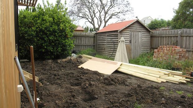 A backyard area under construction, viewed from beside a timber-clad structure. The scene shows freshly excavated soil, stacked timber boards, a sheet of material, gardening tools, a weathered timber shed, timber fencing, and a small stack of bricks. Mature shrubs and trees surround the work area.