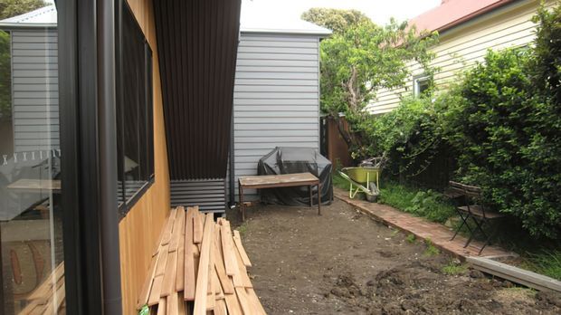 A narrow residential side courtyard or outdoor work area between buildings. The left side shows a contemporary timber-clad structure with dark-framed glazing and a small timber deck edge, while the rear has grey horizontal cladding. The space contains stacked timber boards, a covered outdoor bench, a small side table, garden seating, dense hedging, and a paved strip along the right boundary.