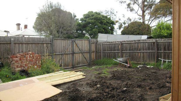 A fenced residential backyard under construction, with freshly excavated soil, stacked timber boards and sheet materials, a wheelbarrow, and a small section of exposed brickwork. Weathered timber fencing and neighbouring rooflines surround the site, with trees visible beyond.