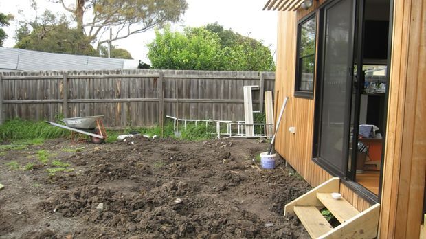 A small backyard beside a timber-clad building, with freshly disturbed soil, a tall timber boundary fence, garden tools, a wheelbarrow, and large black-framed glazed doors and windows. The scene appears to show an outdoor area under landscaping or construction.