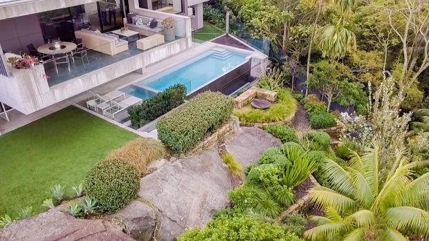 Elevated aerial view of a contemporary residential property with a white open-plan living and dining area, an expansive terrace, and a rectangular swimming pool. The landscaped grounds feature manicured hedges, ornamental planting, natural rock outcrops, palms, and a manicured lawn.