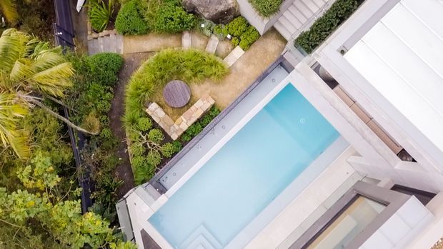 Aerial view of a contemporary residential property showing a rectangular swimming pool bordered by pale paving and light-coloured building surfaces, surrounded by lush tropical and landscaped garden planting, curved garden paths, and outdoor seating.
