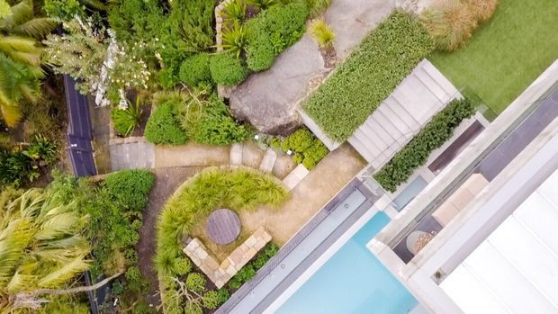 Aerial view of a landscaped residential property with winding stone and concrete garden paths, dense ornamental planting, timber seating, and a bright blue swimming pool beside a modern light-coloured building.
