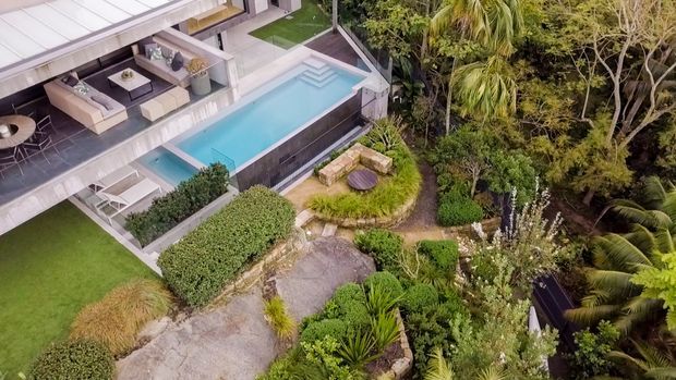 Aerial view of a modern multi-level residence with a rooftop or upper terrace, an elongated swimming pool, outdoor lounge areas, manicured lawn, stone pathways, and dense tropical landscaping. The image is dominated by the landscaped exterior and pool setting.