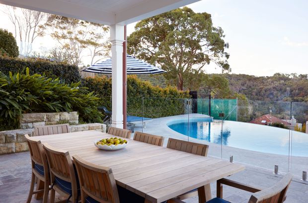 A covered outdoor dining terrace overlooks a glass-fenced swimming pool and wooded suburban landscape. The space features a large timber dining table with matching chairs, a stone-edged garden bed, a striped outdoor umbrella, and a light-coloured rendered column supporting the terrace roof.