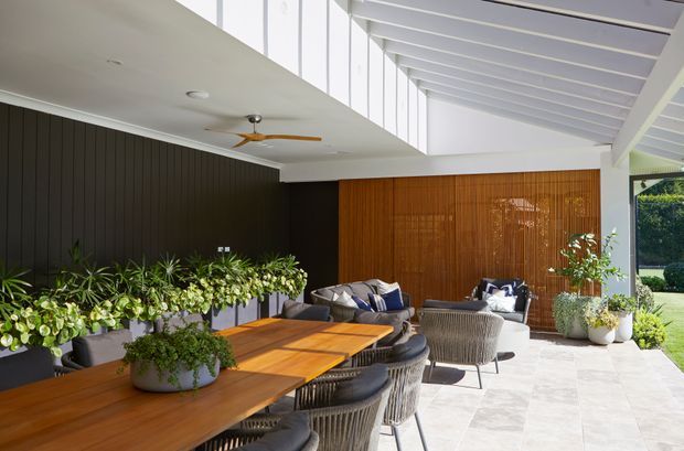 Bright covered outdoor entertaining area with a long timber dining table, woven dining chairs, lounge seating, abundant potted plants, and a striking timber batten feature wall. The space has a white exposed-beam ceiling, high clerestory glazing, and pale stone or concrete-style paving, creating a refined indoor-outdoor living environment.