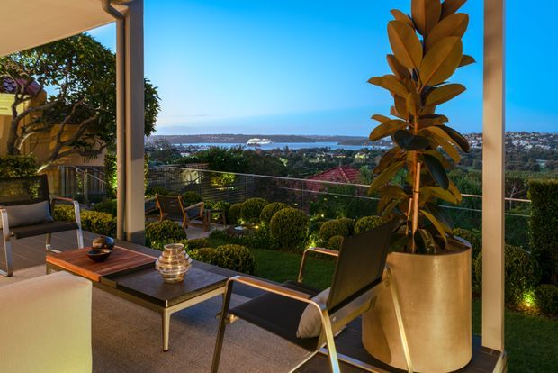 A sophisticated covered outdoor terrace overlooking a wide waterway and distant shoreline. The space features contemporary lounge chairs, a low outdoor coffee table, sculpted hedge planting, integrated garden lighting, and a tall potted plant, creating a refined entertaining area with expansive views.