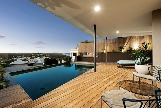 Contemporary outdoor entertaining area with a timber deck, infinity-edge swimming pool, covered terrace, minimalist outdoor furniture, tropical planting, and a landscaped privacy screen. The elevated setting overlooks a suburban landscape at dusk.