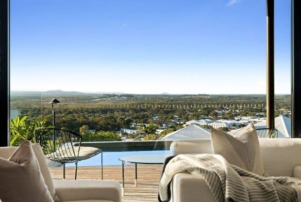 A bright contemporary living area opening onto a timber deck with a narrow pool or water feature. Neutral upholstered seating, a black wire-frame chair, glass coffee table, and soft furnishings frame expansive glazing and panoramic views over a landscaped semi-rural landscape and distant hills.