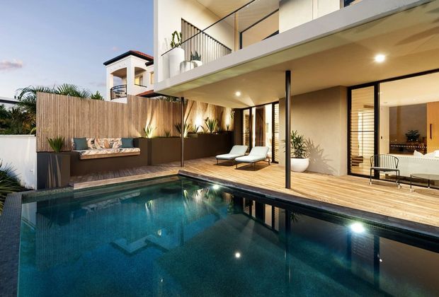 Modern multi-level residential courtyard viewed from the pool, featuring a dark tiled swimming pool, timber decking, a covered outdoor entertaining area, built-in bench seating, bamboo screening, lush planting, and extensive glazed doors opening to the interior. The upper level has a balcony with slim black railings and a clean contemporary facade.