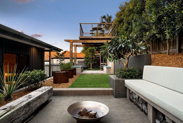 A contemporary landscaped courtyard with stepped concrete paving, built-in outdoor seating, raised garden beds, mature planting, and a compact lawn. A timber-framed elevated deck or terrace with open balustrading and a stairway forms a strong architectural feature at the rear. Dark cladding, warm timber, rendered walls, and terracotta roof tiles are visible around the courtyard.
