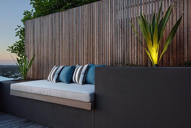 A contemporary outdoor lounge or terrace with a built-in dark rendered bench, pale upholstered seat cushion, blue and striped cushions, tall timber privacy screening, and tropical architectural plants. The setting appears elevated, with a distant coastal or urban view beyond the screen.