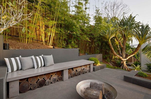 A landscaped outdoor courtyard or terrace featuring a long built-in concrete bench with a decorative stacked-log base, striped and grey cushions, and a round fire bowl in the foreground. Dense bamboo screening and tropical sculptural planting create a private, resort-like setting, with dark rendered planter walls and paved flooring.