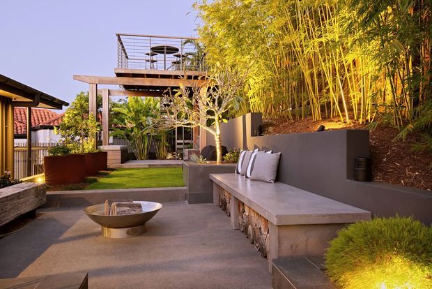 A landscaped outdoor courtyard or terrace with a built-in concrete bench, low fire bowl, paved flooring, tropical planting, bamboo screening, and a timber pergola structure with an elevated deck or roof terrace. The space has a warm, contemporary resort-like feel and connects to adjoining residential structures.