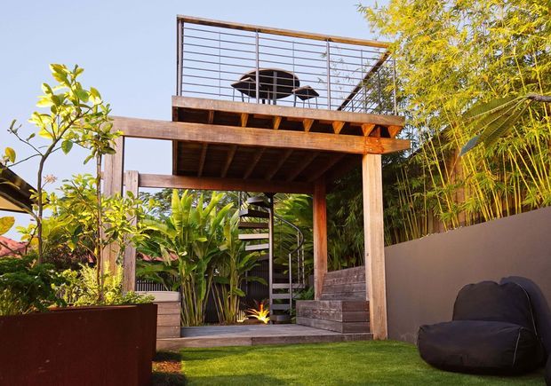 A tropical landscaped outdoor area featuring a raised timber deck and rooftop terrace, supported by robust timber posts and beams. A spiral staircase connects the lawn-level garden to the upper deck, which has horizontal metal railing and outdoor furniture. Dense tropical planting, bamboo screening, a rendered boundary wall, planter boxes, and a lawn create a lush courtyard-like setting.
